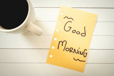 Üzerinde günaydın yazan bir not ve bir fincan kahve. Beyaz ahşap arka planda.