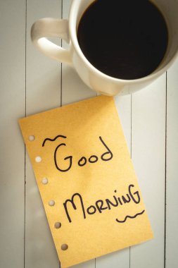 Üzerinde günaydın yazan bir not ve bir fincan kahve. Beyaz ahşap arka planda.