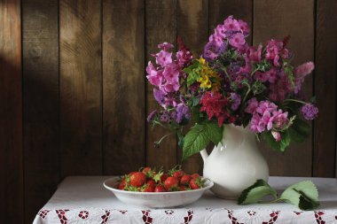 Masanın üzerinde pembe Phlox buketi ve beyaz sürahi içinde mor ageratum ve çilek. Kırsal bir iç mekanda kompozisyon.
