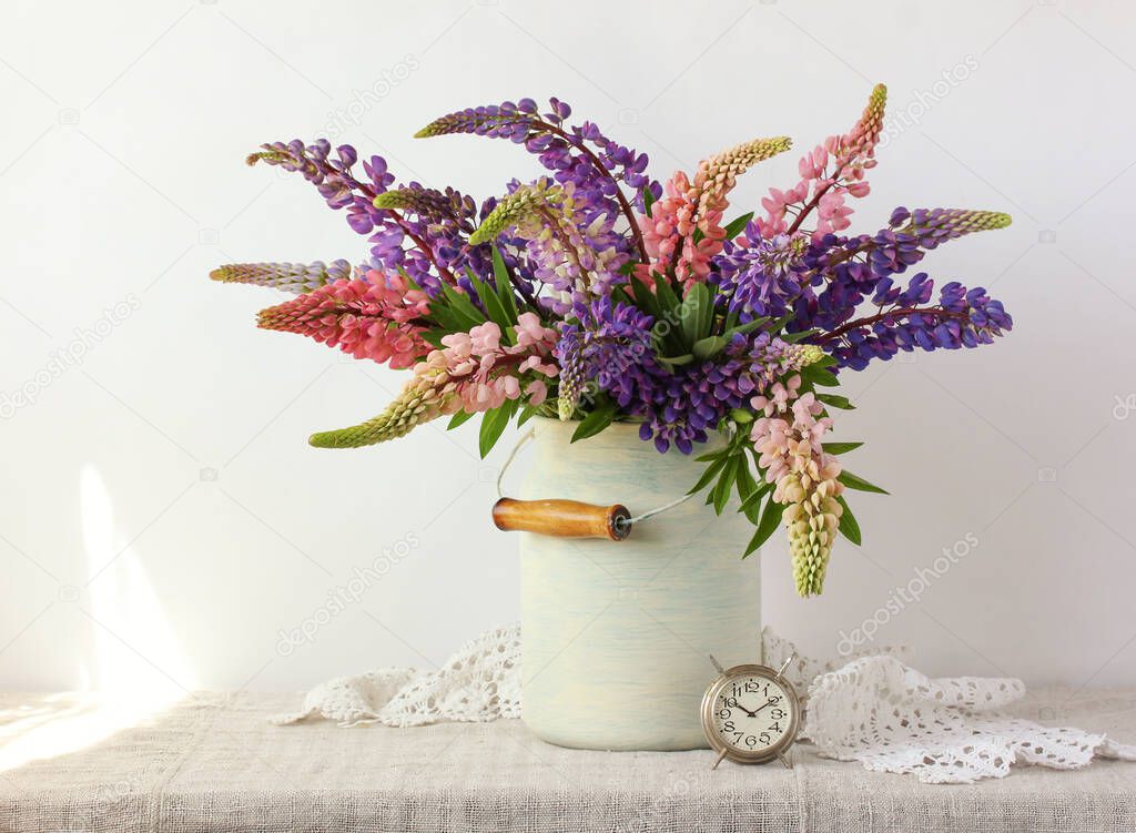 summer composition with a bouquet of lupines and a metal alarm clock on a light background.