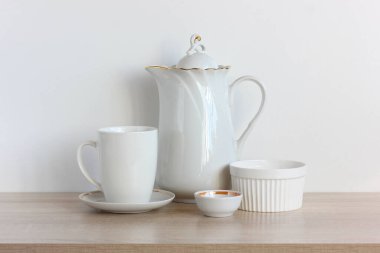 Masada bir grup beyaz tabak var. Mutfak aletleri rafta. Kahve demliği ve kupa..