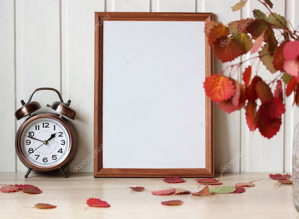 autumn mockup with a bouquet of leaves and an empty wooden frame on the table September natural background interior with an alarm clock
