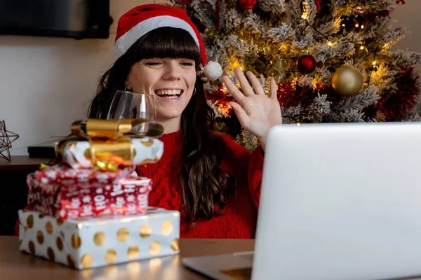 Young girl in Christmas hat and red sweater making a video call to her family to show them Christmas gifts while having a glass of wine with the Christmas tree behind