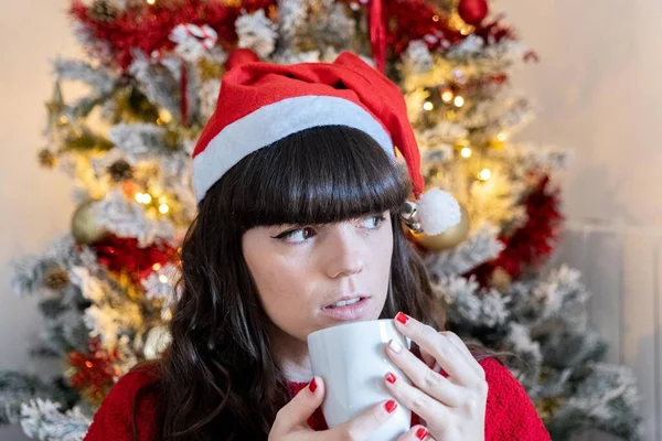 Young girl in Christmas hat and red sweater enjoying a coffee in front of the Christmas tree