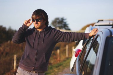 Bir yere bakan güneş gözlüklü, park halindeki bir arabaya yaslanan genç adam.