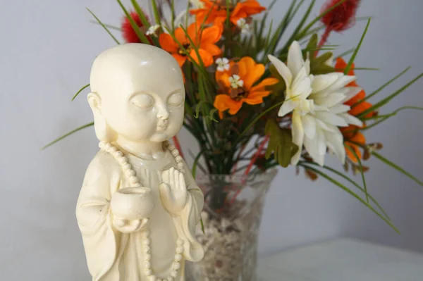 İçinde renkli çiçekler olan cam bir vazonun yanında beyaz bir Buda figürü. Beyaz bir duvarın yanındaki beyaz ahşap bir masanın üzerine yerleştirilmiş. İçeriye doğal aydınlatmayla götürüldü..