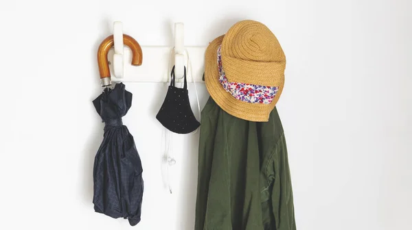 things you need if you are going outside in the new normal because of covid-19. Hanging from a coat rack an umbrella, a coat, face masks and headphones. Over a white wall