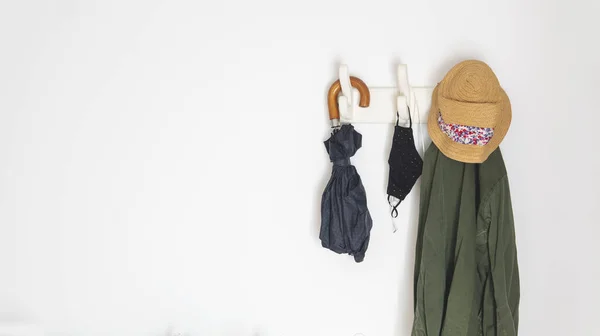 things you need if you are going outside in the new normal because of covid-19. Hanging from a coat rack an umbrella, a coat, face masks and headphones. Over a white wall