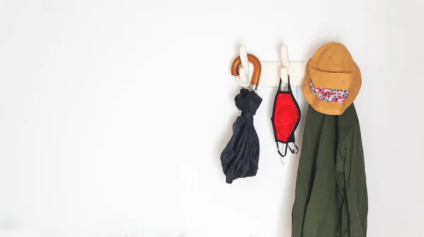 things you need if you are going outside in the new normal because of covid-19. Hanging from a coat rack an umbrella, a coat, face masks and headphones. Over a white wall