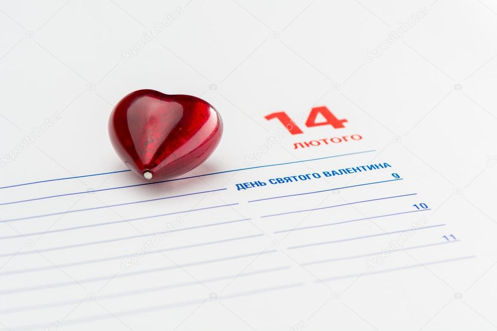 Red heart lies on a page of Diary. Diary is opened at 14 February page.  Text on the Diary page is written in ukrainian and means 14 February,  st. Valentines day.