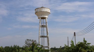Kulede beyaz beton su tankı. Toplumlar ya da işletmeler için su deposu. Aşağıda elektrik kabloları ve yeşil ağaçlar var. Gökyüzü arka planında kopyalama alanı olan beyaz bulutlar var.