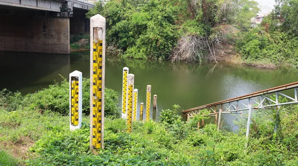 Pillar water level gauge.Yellow water level plate on many concrete poles on the shore for river volumetric measurement on water background, bridges and green trees with copy area. Selective focus