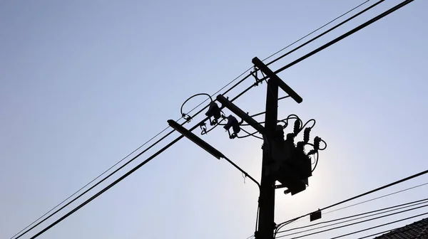 Silhouette of a transformer on a pole. Three-phase distribution transformer for converting high voltage with transmission lines on insulators and street lamps in a sunny sky background with copy space.