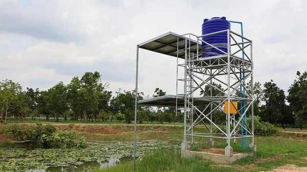 Metal structure of the blue water tank. Water tank with solar panels in farm groundwater pumping system on green tree background and white cloudy sky with copy space. Selective focus