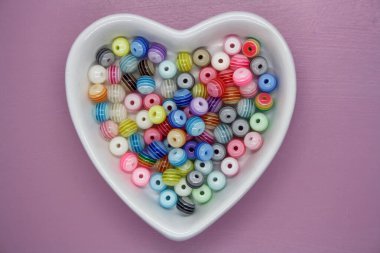 Pembe arka planda kalp şeklinde bir kasede renkli boncuklar..