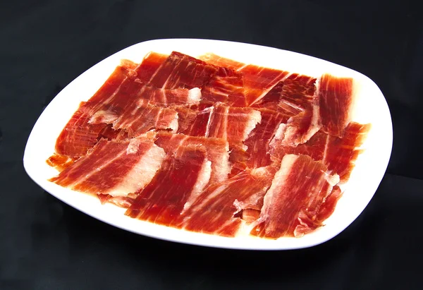 Serrano ham slices on a white dish over black wackground. Jabugo. Spanish tapa. — Stock Photo, Image