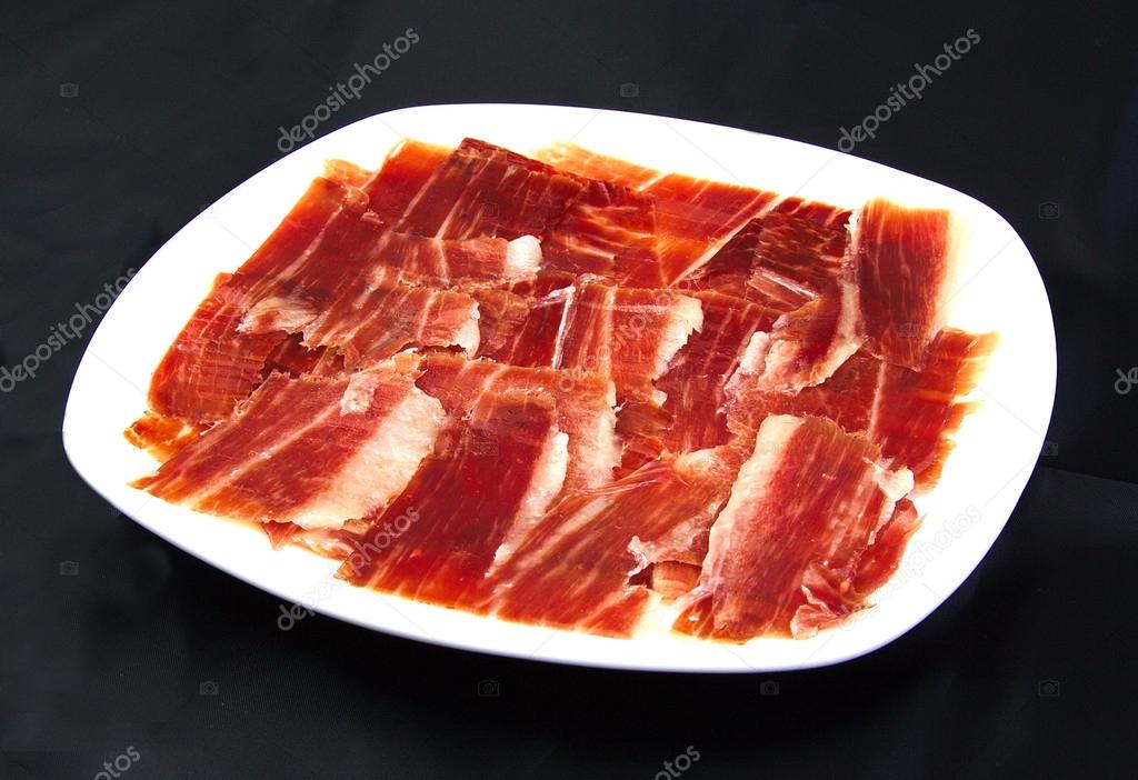 Serrano ham slices on a white dish over black wackground. Jabugo. Spanish tapa.
