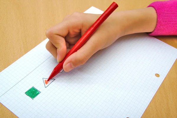Criança desenhando um triângulo vermelho e um quadrado verde com um lápis de cera vermelha . — Fotografia de Stock