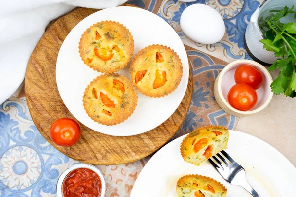 Small egg bite pastry muffins on a handmade white ceramic plate with a white linen napkin and egg, tomato, on a wooden board, and tile background. Finger food and mini food concept. Overhead. Top View