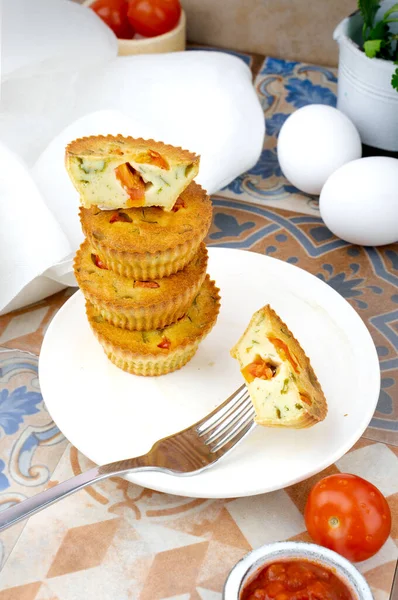 Small egg bite pastry muffins on a handmade white ceramic plate with a white linen napkin and egg, tomato, on a wooden board, and tile background. Finger food and mini food concept. Overhead. Top View