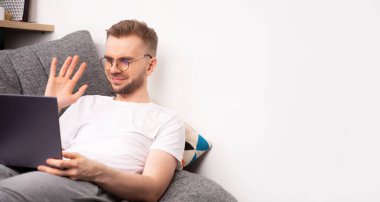 İnternetteki bir öğretmenden, okuldan öğreniyor. Uygulamanın sosyal ağı üzerinden arkadaşlarınızla web kamerasından iletişim kurun. Sarışın bir adam evde kanepede oturuyor ve ekrana el sallıyor. Uzayı kopyala Pankart.