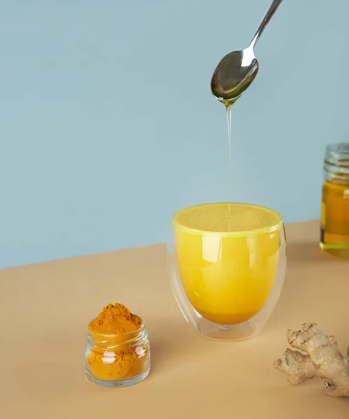 Golden yellow milk with turmeric in a glass on with ginger and honey in spoon on a isometric diagonal projection blue and beige background.healthy natural energy drink.Concept turmeric latte.Vertical.