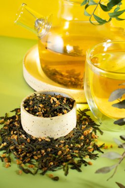 Japon yeşil çayı Genmaicha.Çay yaprakları ve kızarmış kahverengi pirinç, parlak sarı arka planda gölgeli, trend çay konsepti. Bir fincan çay. Şeffaf bir çaydanlık demle