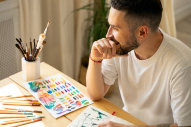 Stüdyoda güneş ışığıyla aydınlatılan beyaz tişörtlü, beyaz sakallı, siyah saçlı beyaz erkek ressamın portresi. Ahşap bir masada renkli kalemler ve boya fırçaları. Yaşam tarzı. Modern oda