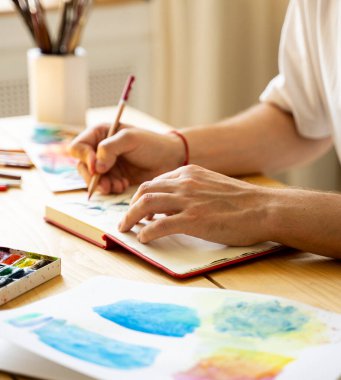 Genç bir sanatçı ahşap bir masada otururken bir defter çizer. Güzel erkek eller renkli bir kalem tutuyor. Yakın plan. Güneş ışınları ve hoş bir atmosfer. Konsept diy. Gerçek bir sanat eseri.