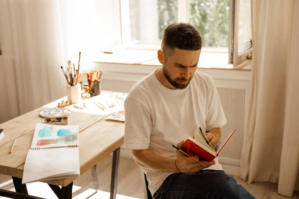 Portrait of Young dark haired concentrated caucasian white male artist with a beard in a white t-shirt. sunlight. sitting in a studio in wooden table.in hand holding colored pencil.diy.