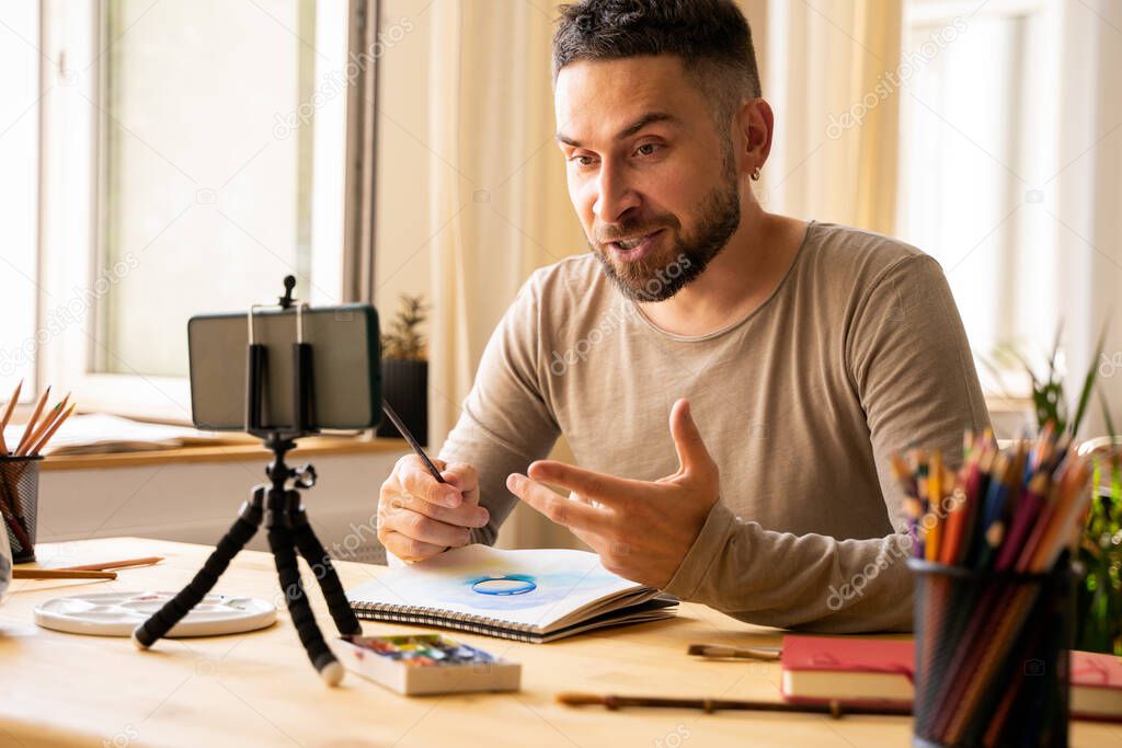 Portrait of Mid-adult artist male. Dark haired man smiling happy making online art class for students at studio. Holding watercolor drawing in his hand. Concept zoom Remote learning. Real emotions.