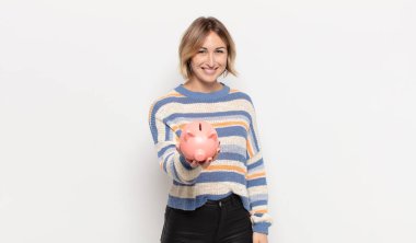 Genç sarışın kadın mutlu bir şekilde gülümsüyor, eli kalçasında ve kendinden emin, pozitif, gururlu ve arkadaş canlısı.