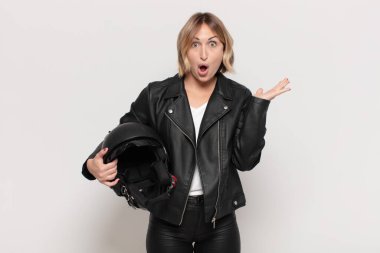 Genç sarışın kadın şaşırmış ve şoka uğramış görünüyor. Çenesi düşmüş ve eli açık bir objeyi tutuyor.