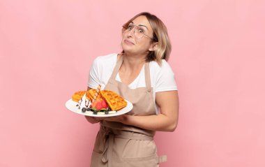 Genç sarışın kadın omuz silkiyor, kafası karışık ve kararsız, kolları bağlı ve şaşkın görünüyor.