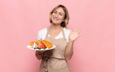 Genç sarışın kadın mutlu ve neşeyle gülümsüyor, el sallıyor, seni selamlıyor ve selamlıyor, ya da hoşça kal diyor.