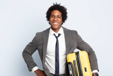 Genç siyah afro adam mutlu bir şekilde gülümsüyor, eli kalçasında ve kendinden emin, olumlu, gururlu ve arkadaş canlısı.