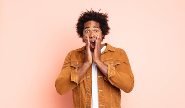 Genç siyah afro adam şok olmuş ve korkmuş hissediyor, ağzı açık ve elleri yanaklarında korkmuş görünüyor.