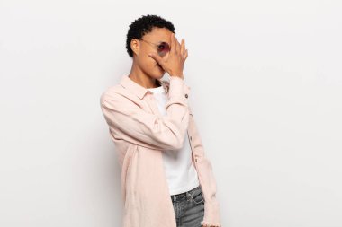 Genç siyah afro kadın şok olmuş, korkmuş ya da korkmuş görünüyor. Yüzü elle kaplanmış ve parmaklarının arasından gizlice bakıyor.