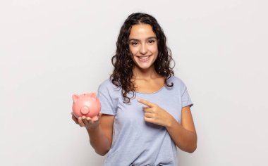 Genç İspanyol kadın neşeyle gülümsüyor, mutlu hissediyor ve kenarı ve yukarıyı işaret ediyor, fotokopi alanında nesneler gösteriyor.