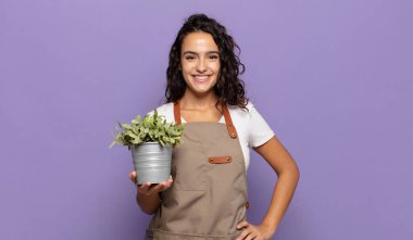 Genç İspanyol kadın mutlu bir şekilde gülümsüyor, eli kalçasında ve kendinden emin, olumlu, gururlu ve arkadaş canlısı bir tavırla.