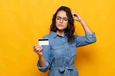 Genç İspanyol kadın şaşkın ve şaşkın hissediyor, kafasını kaşıyor ve yan tarafa bakıyor.