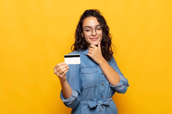 молодая латиноамериканка улыбается со счастливым, уверенным выражением лица с рукой на подбородке, удивляясь и глядя в сторону