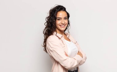 Çapraz kollarla kameraya gülümseyen güzel genç bir kadın ve mutlu, kendinden emin, tatmin olmuş bir ifade, yan görüş.