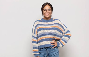 Güzel, genç bir kadın mutlu bir şekilde gülümsüyor, eli kalçasında ve kendinden emin, olumlu, gururlu ve arkadaş canlısı bir tavırla.