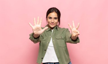 Güzel genç bir bayan gülümsüyor ve arkadaş canlısı görünüyor, 8 ya da 8 numarayı gösteriyor, elleri önde, geri sayıyor.