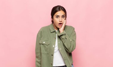 Güzel genç bir kadın şok ve inançsızlık içinde, elleri yanak ve kolları çaprazlanmış, sersemlemiş ve şaşırmış hissediyor.