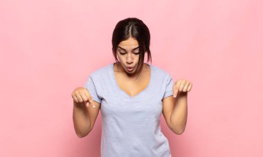 Ağzı açık, iki eliyle aşağıya doğru bakan güzel genç bir kadın. Şok olmuş, şaşırmış ve şaşırmış görünüyor.