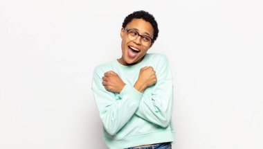 Genç afro kadın sevinçle gülümsüyor ve kutluyor, yumruklarını sıkıp kollarını kavuşturuyor, mutlu ve pozitif hissediyor.