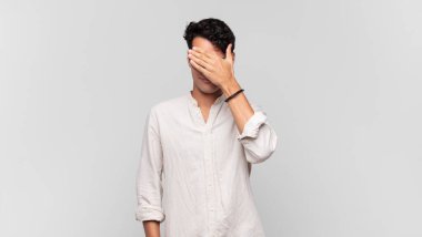 Genç ve yakışıklı bir adam stresli, utanmış ya da üzgün görünüyor, baş ağrısı var, yüzünü eliyle kapatıyor.