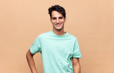 Genç yakışıklı adam mutlu bir şekilde gülümsüyor, eli kalçasında ve kendinden emin, olumlu, gururlu ve arkadaş canlısı.